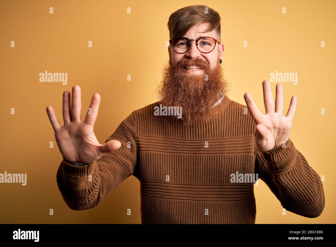 Handsome Irish redhead man with beard wearing glasses and winter sweater over yellow background showing and pointing up with fingers number nine while Stock Photo