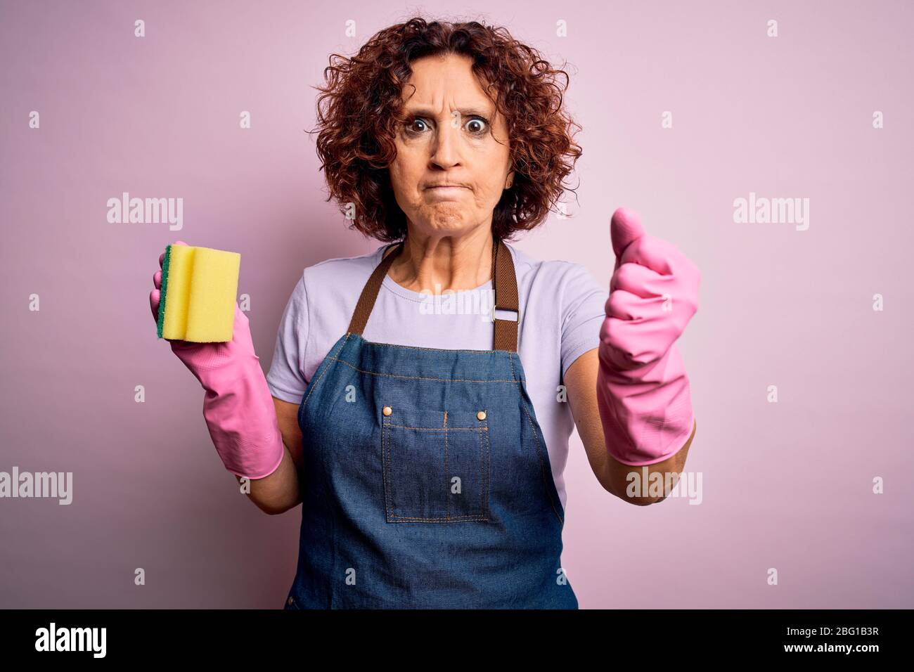 Middle age curly hair woman cleaning doing housework wearing apron and gloves using scourer annoyed and frustrated shouting with anger, crazy and yell Stock Photo