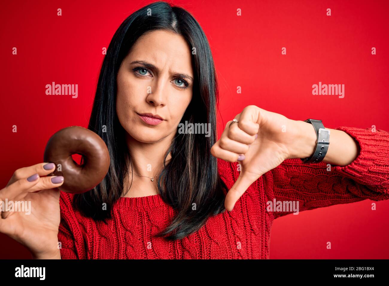 Young brunette woman with blue eyes eating chocolate doughnut over red ...