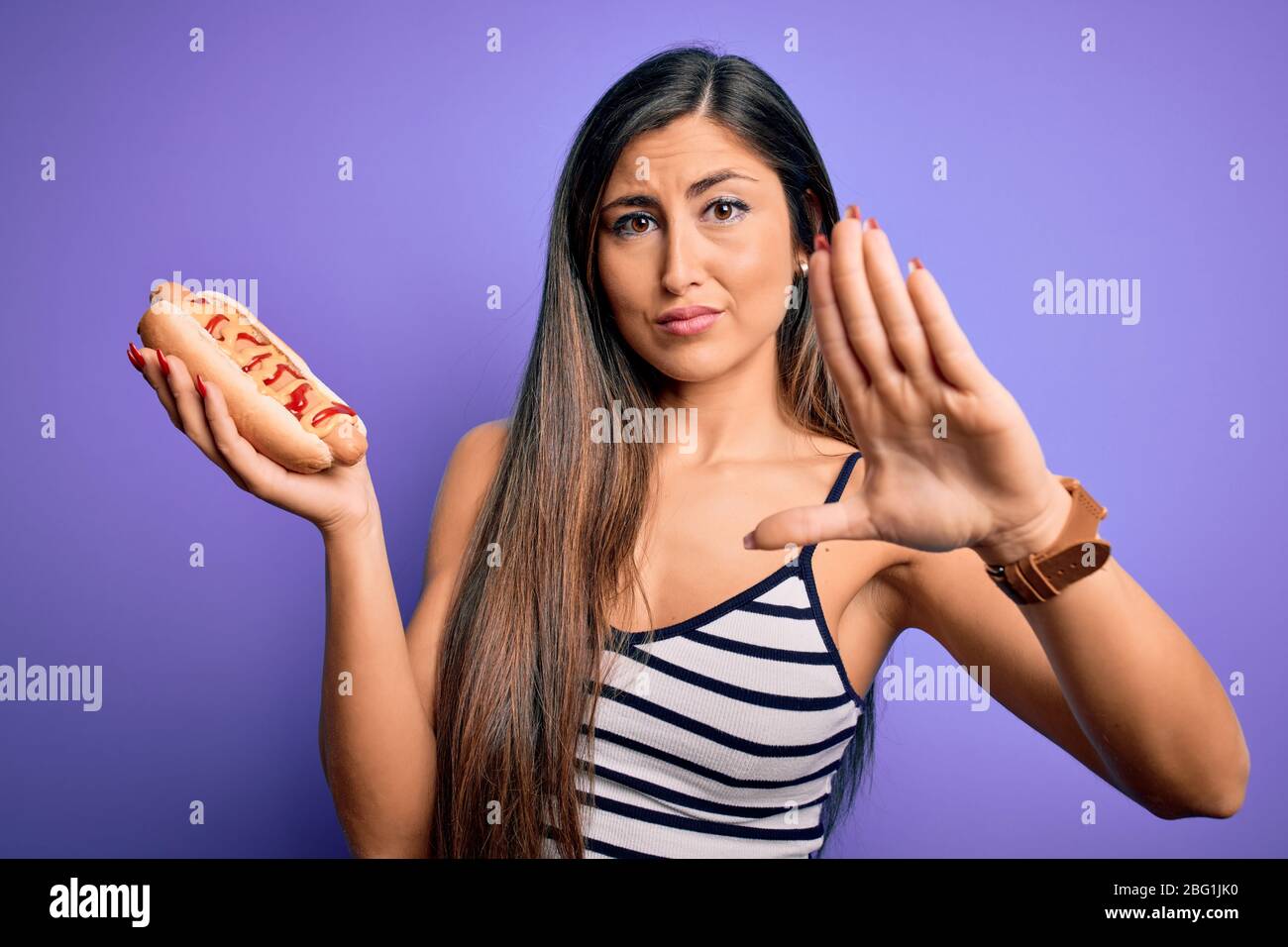 Young woman eating hotdog with ketchup and mustard over purple