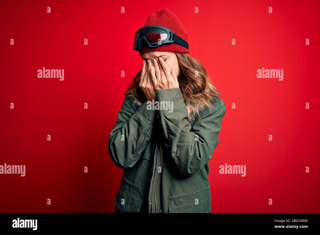 Young blonde girl wearing ski glasses and winter coat for ski weather over red background rubbing eyes for fatigue and headache, sleepy and tired expr Stock Photo