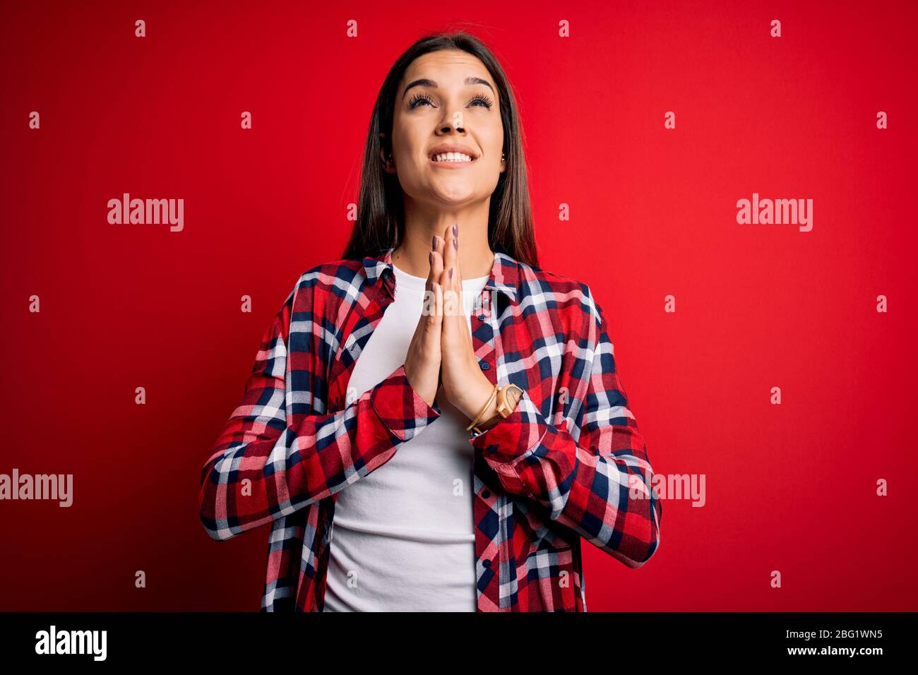 Young beautiful brunette woman wearing casual shirt standing over isolated red background begging and praying with hands together with hope expression Stock Photo