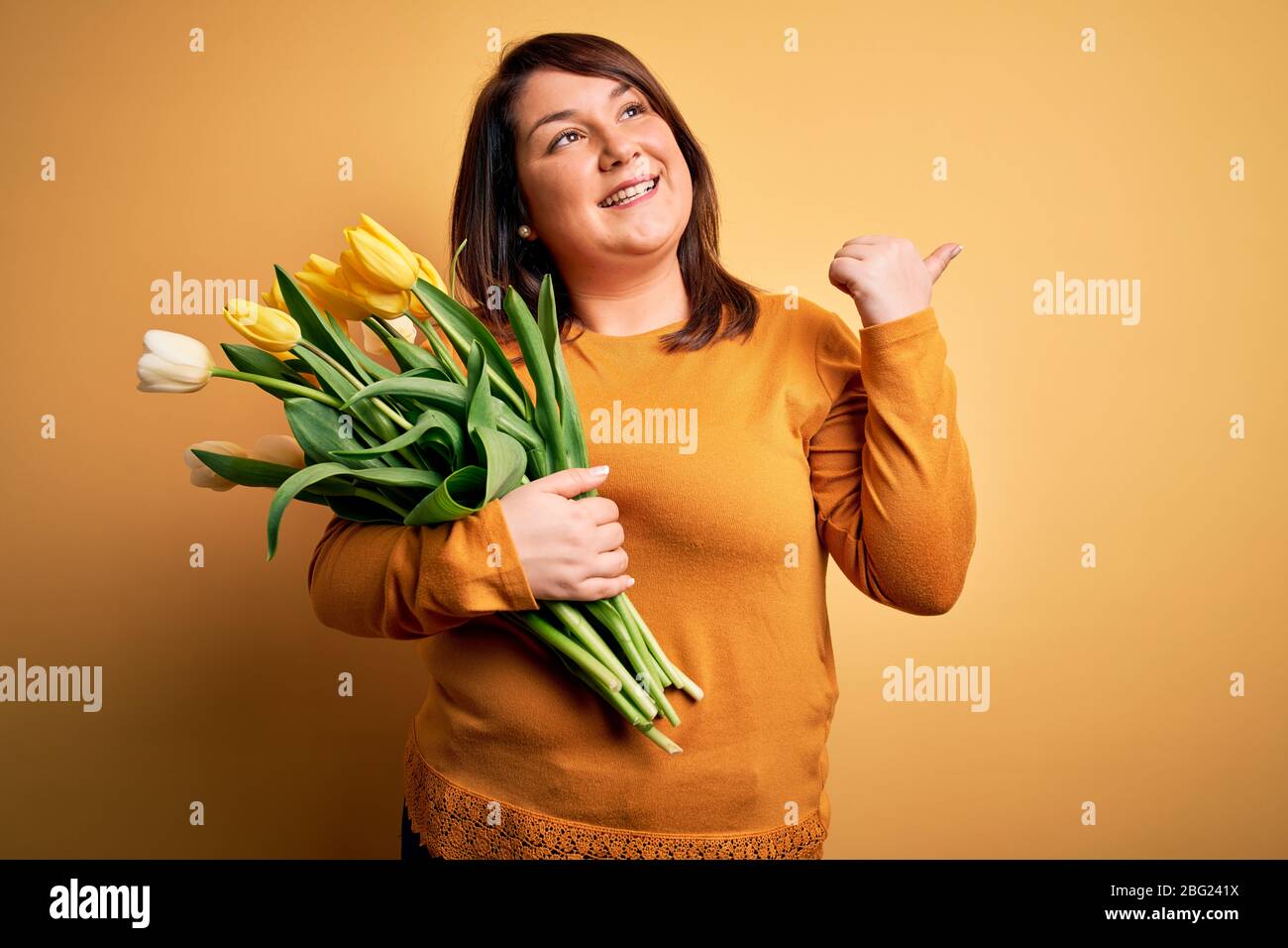 Beautiful plus size woman holding romantic bouquet of natural tulips flowers over yellow background smiling with happy face looking and pointing to th Stock Photo
