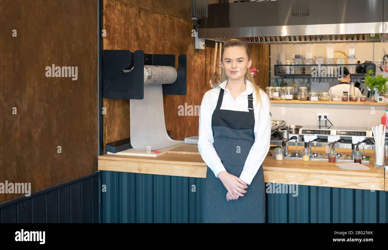 Young waitress wearing apron posing in restaurant. Portrait of a young woman on her first day job in a coffee shop. Smiling young girl or small cafe b Stock Photo