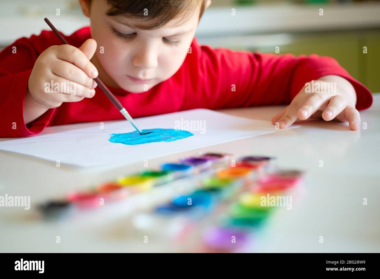 Boy Learns to Paint with a Brush on Paper Sitting at a Table Stock Photo