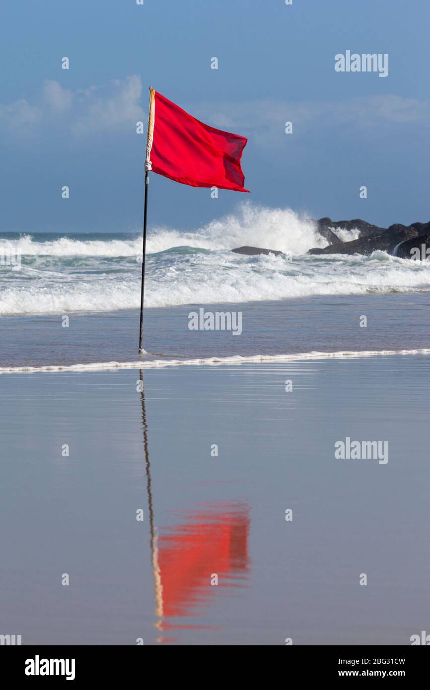 A red Safety flag flapping in the wind with large waves breaking on the rocks behind. Stock Photo