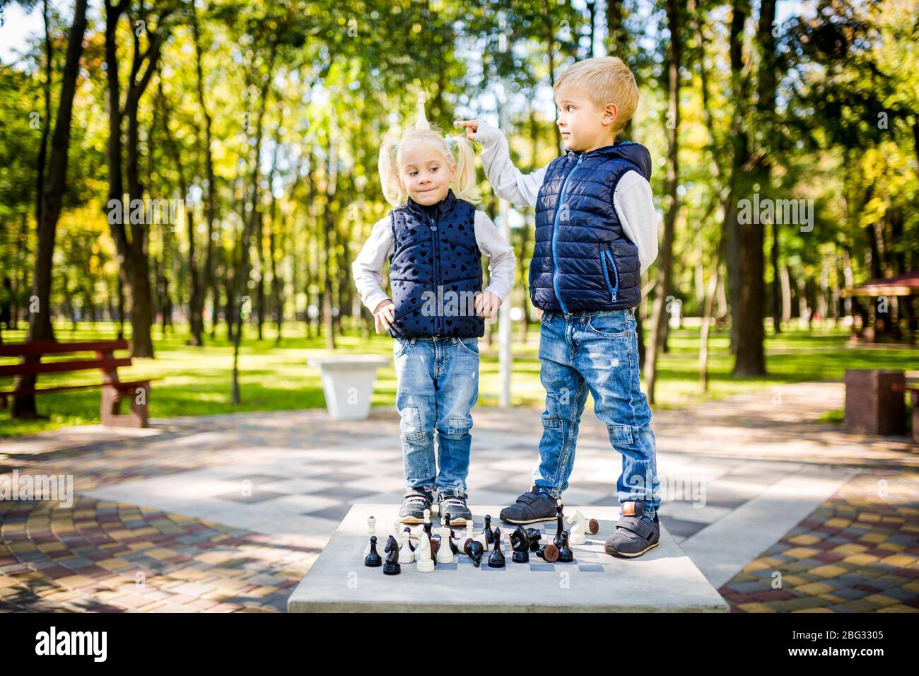 Funny little brother and sister of Caucasian ethnicity play on a public chess venue in a city park. Children frolic and indulge instead of studying Stock Photo