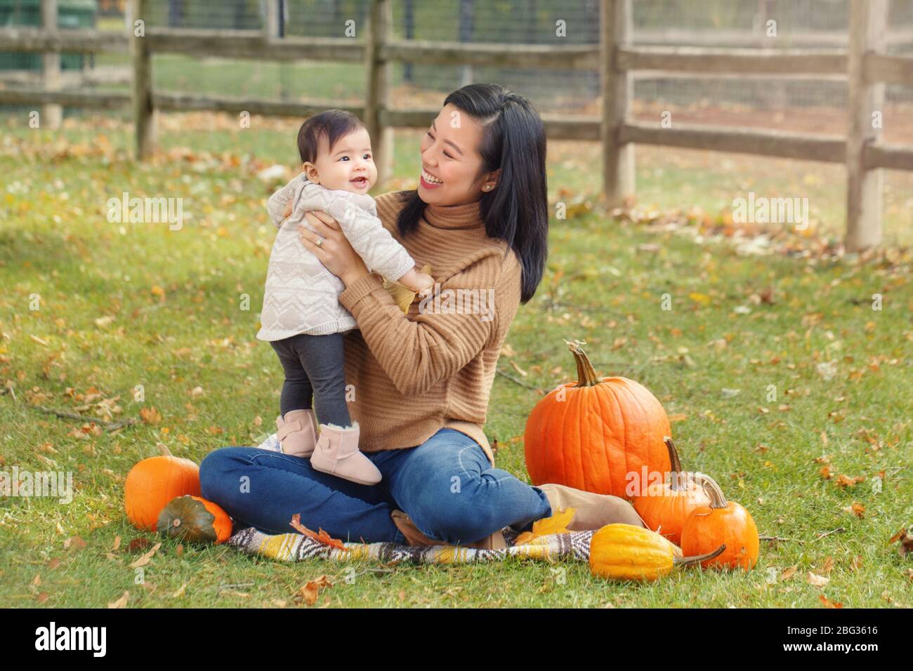 Happy smiling Asian Chinese mother with cute adorable baby. Mom and daughter girl family sitting in autumn fall park outdoor with pumpkins. Halloween Stock Photo