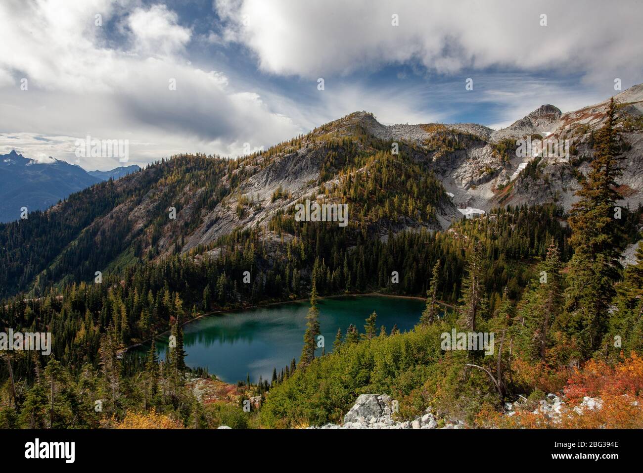 A mountain lake in a basin is visible from the side of a mountain, surrounded by Western larch trees, under a blue sky with white and light grey cloud Stock Photo