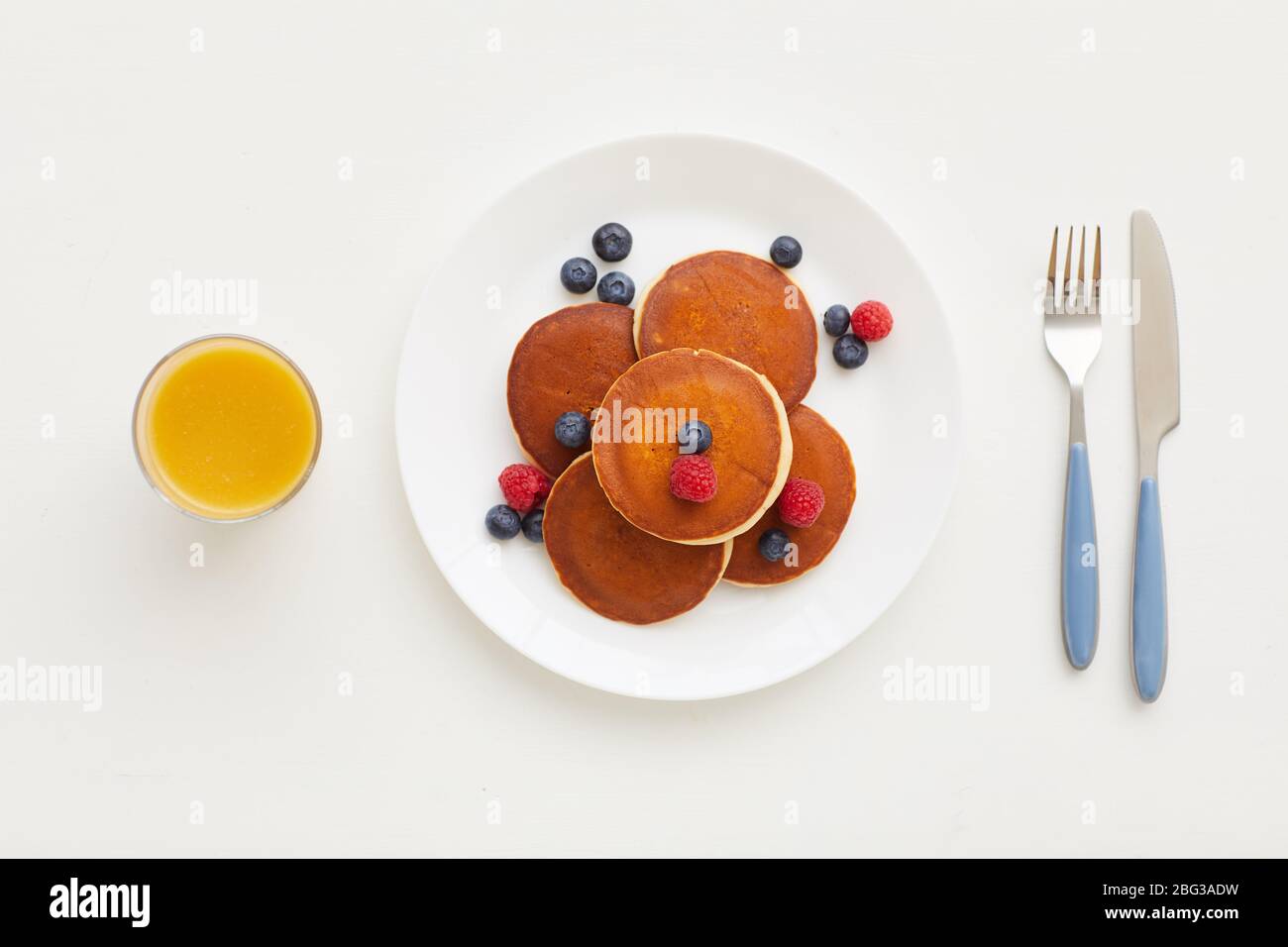 Top view at minimal composition of delicious golden pancakes with fresh berries next to orange juice and knife and fork on white background, breakfast Stock Photo