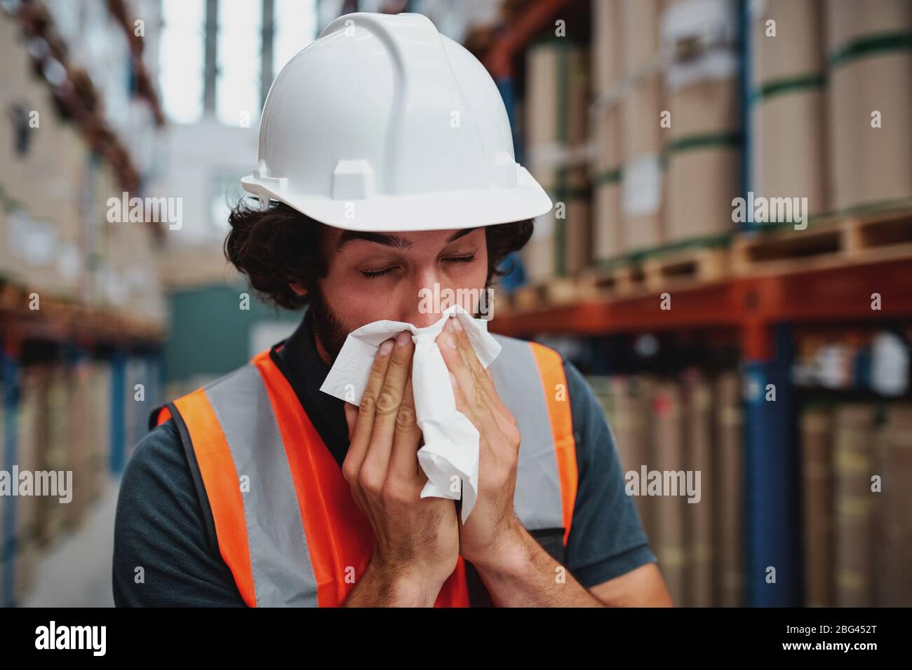 Young warehouse worker feeling sick while sneezing and coughing at factory standing near packed cardboard boxes on shelf Stock Photo