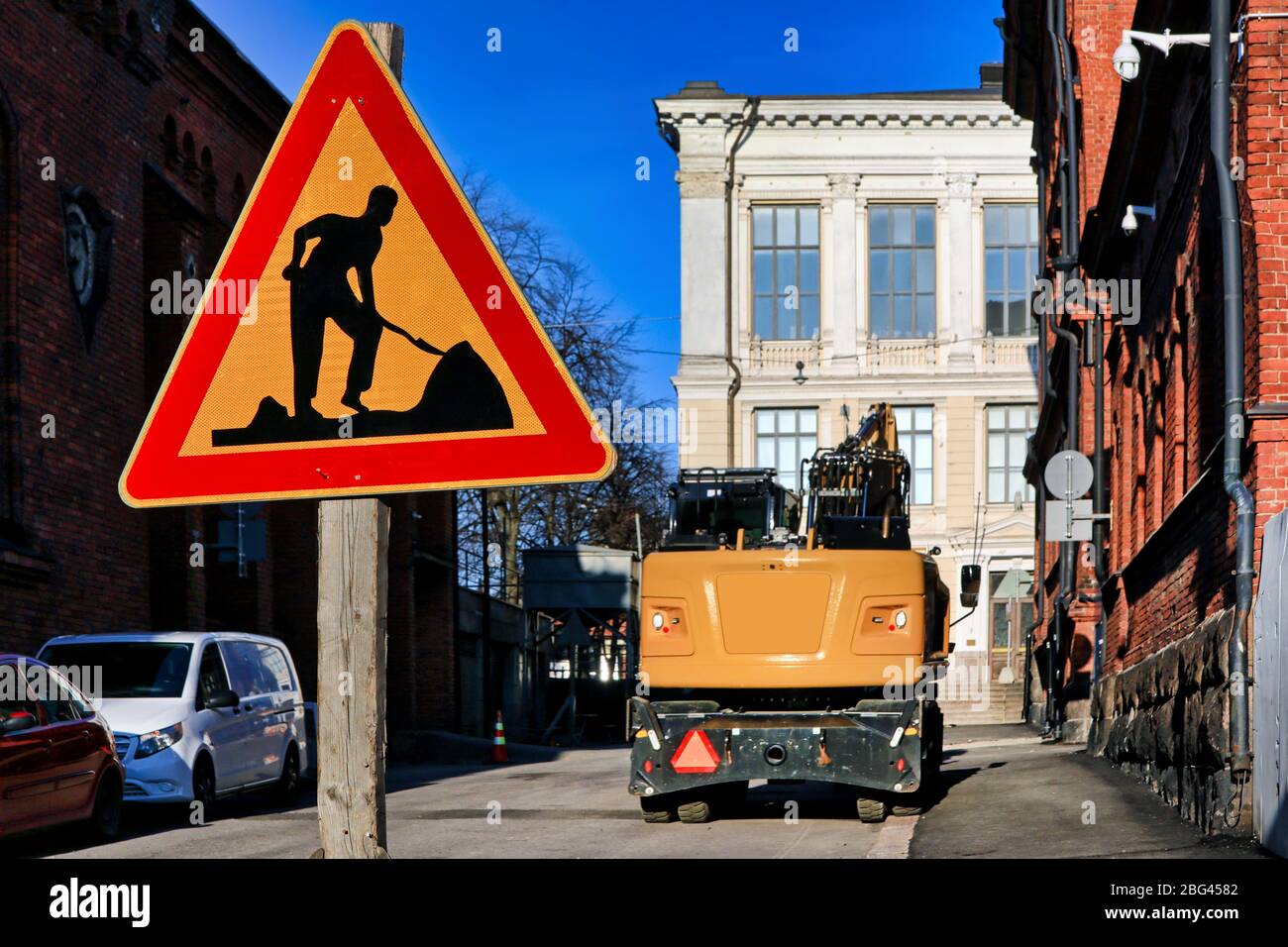 Road works traffic sign by city street on a sunny day with yellow hydraulic excavator on the background. No people, branding removed from image. Stock Photo