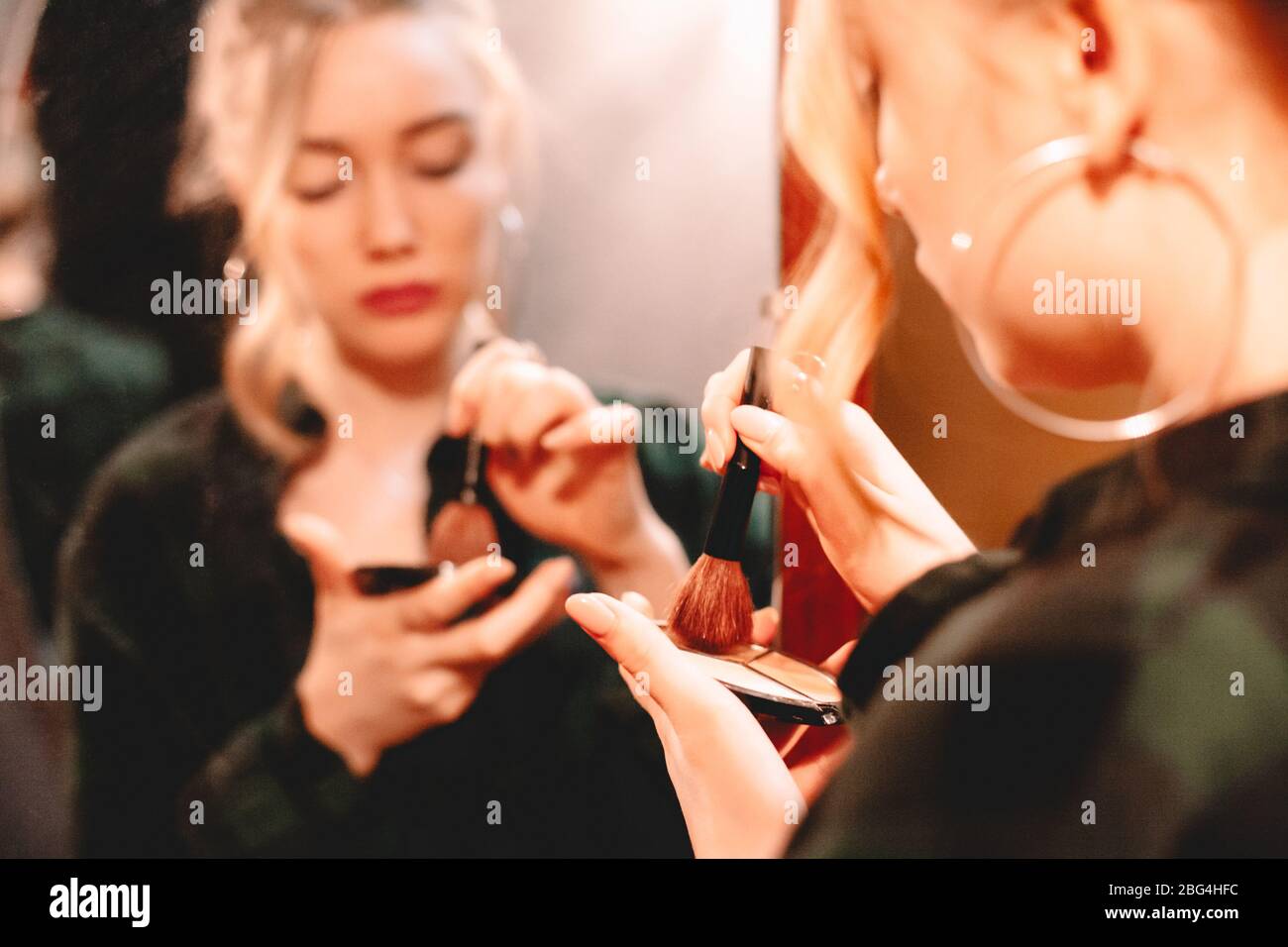 Young woman using palette and makeup brush while applying make up standing in front of mirror at home getting ready to go out Stock Photo