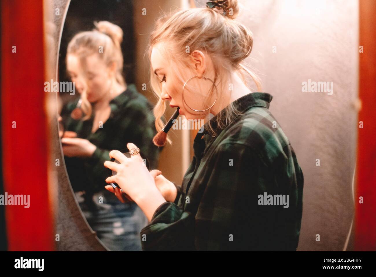 Young woman opening palette and holding makeup brush in her mouth while applying make up standing in front of mirror at home getting ready to go out Stock Photo