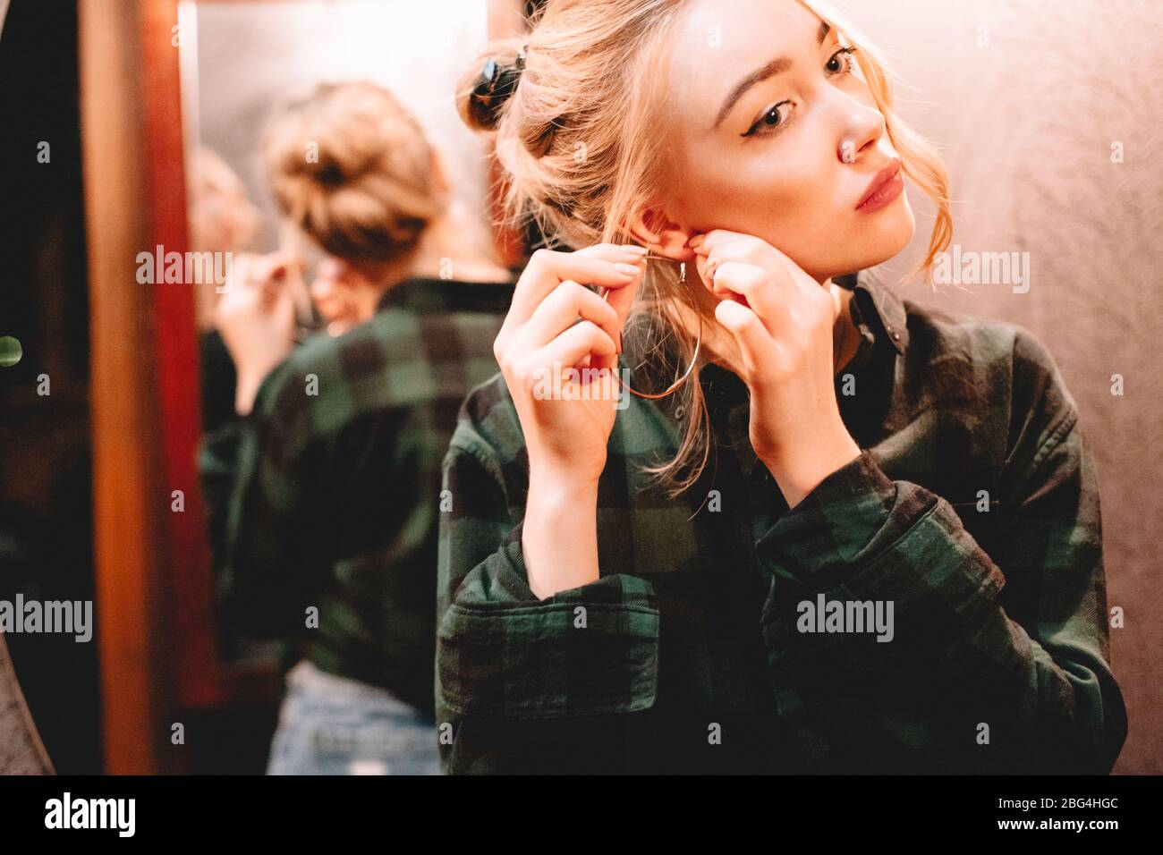 Young woman putting on hoop earring while standing in front of mirror at home getting ready to go out Stock Photo