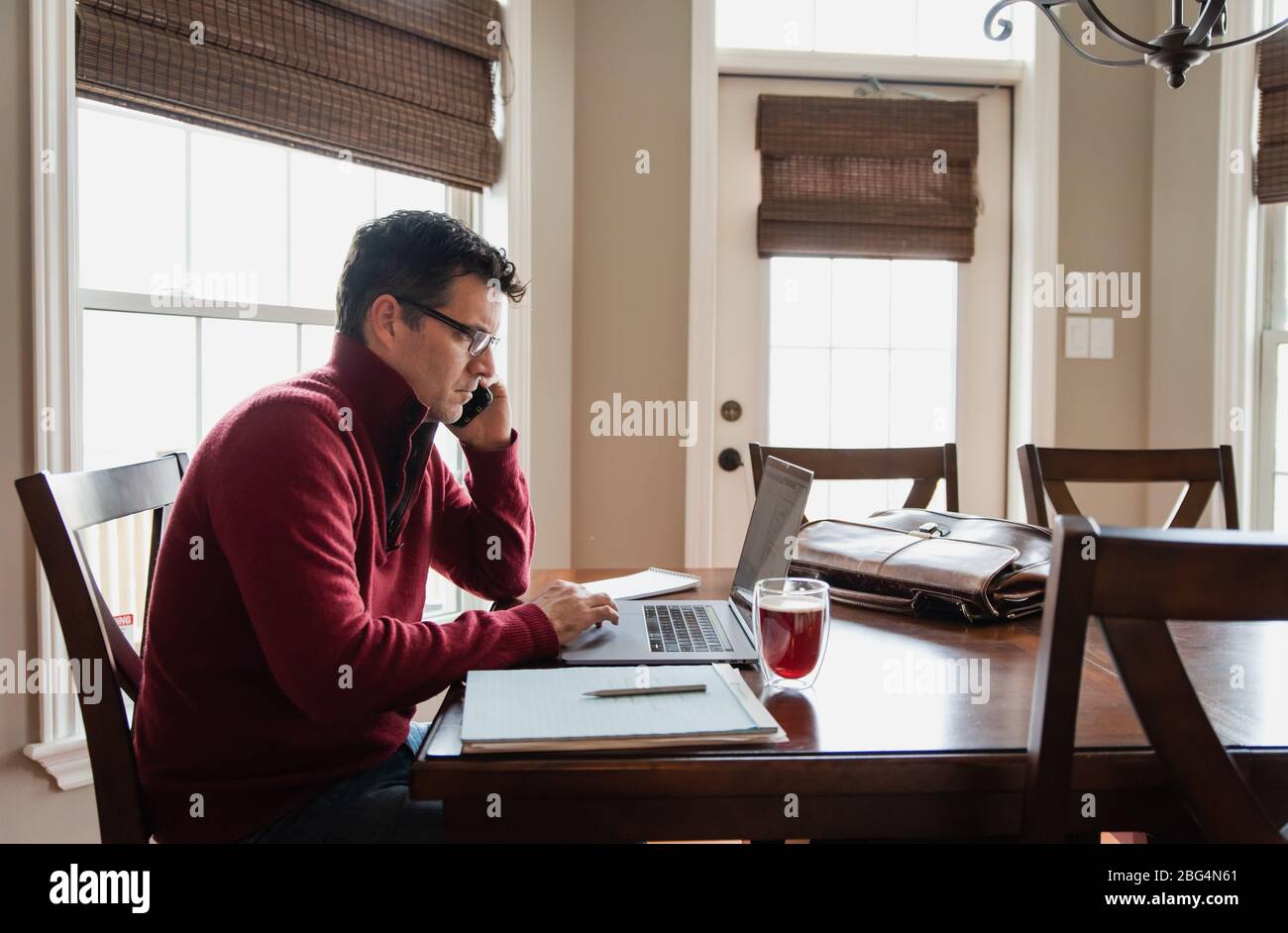 Man on cellphone working from home using a computer at a dining table. Stock Photo
