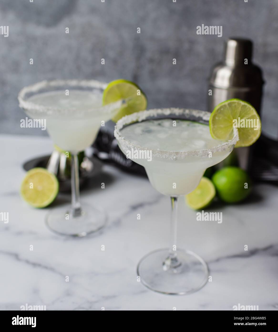 Two margarita cocktails with limes and shaker on white marble counter