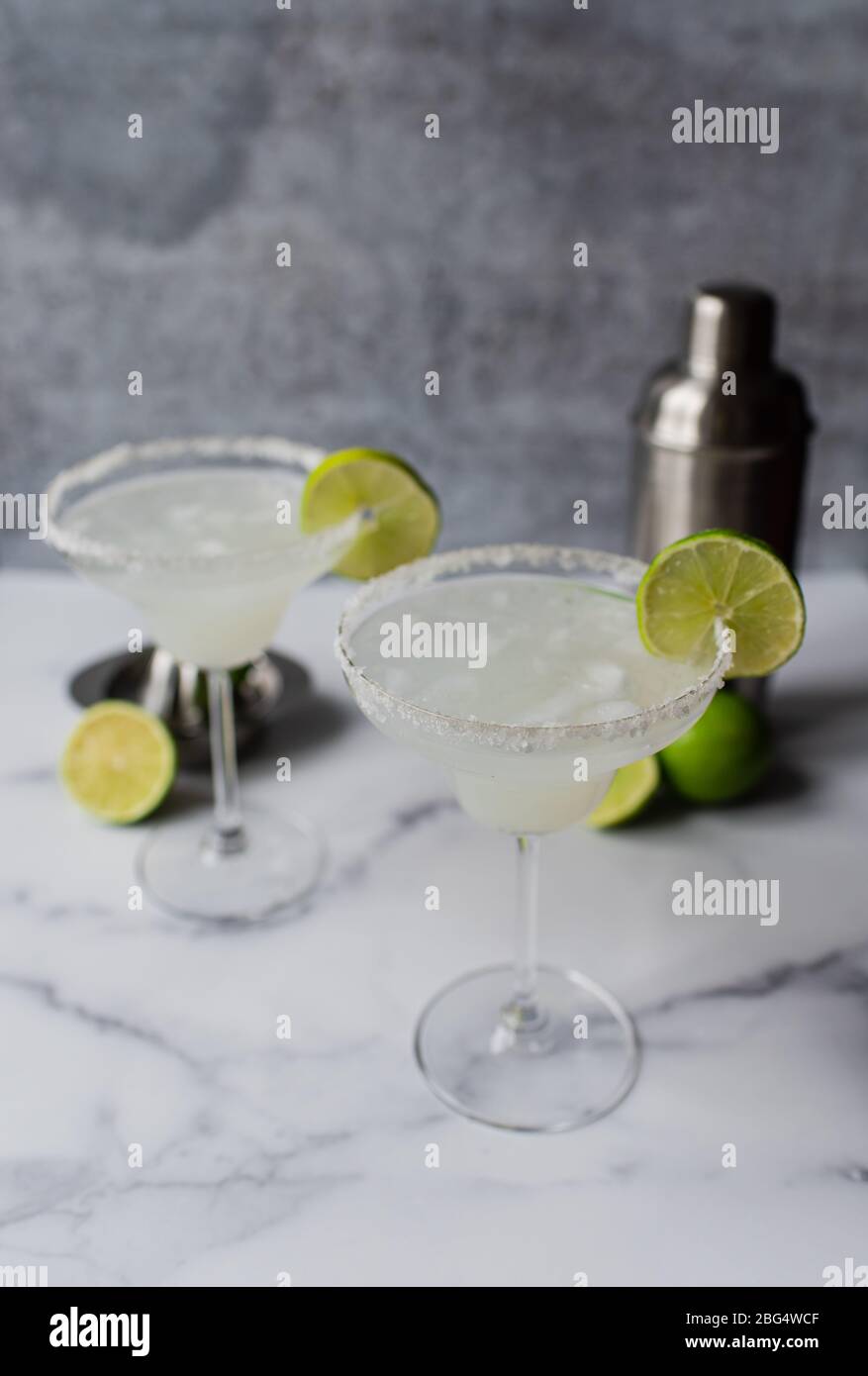 Two margarita cocktails with limes and shaker on white marble counter