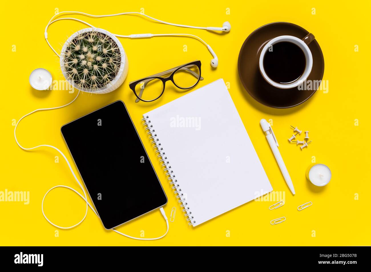 Flat lay of modern home office workspace with tablet, coffee cup, note pad, pen, eyeglasses and cactus on yellow background. Top view Stock Photo