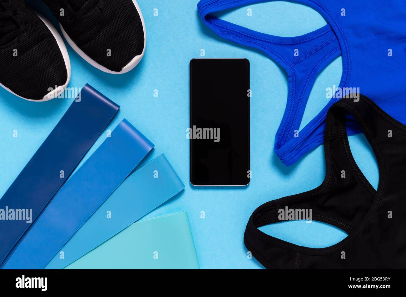 Flat lay of sports bra, running shoes, mobile phone and resistance bands on blue background. Sports equipment for fitness at home. Healthy lifestyle, Stock Photo