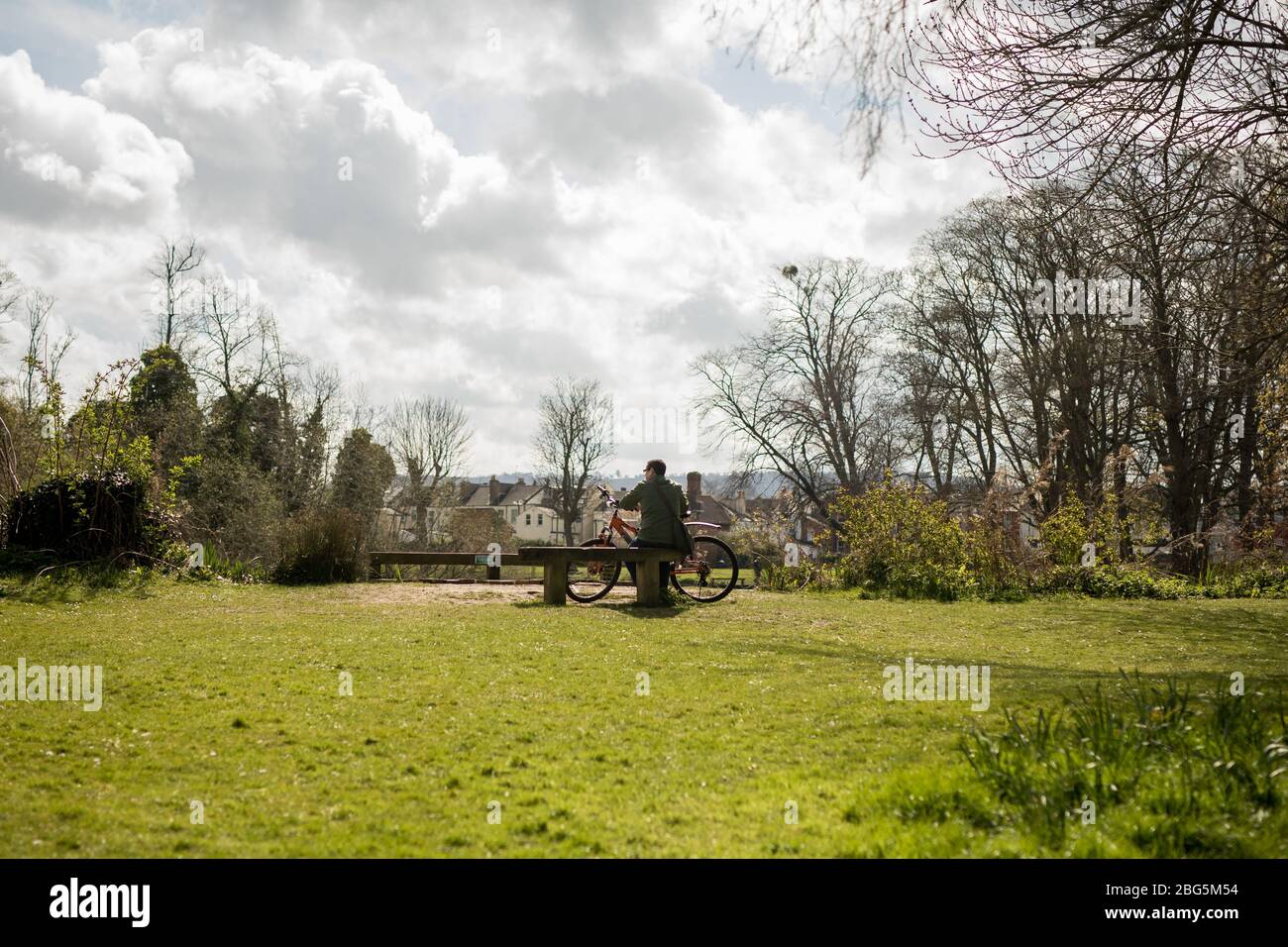 A far off shot of a man sat on a bench surrounded by grass, in front of the lake in Pitville Park, Cheltenham with his bike on a bright cloudy day Stock Photo