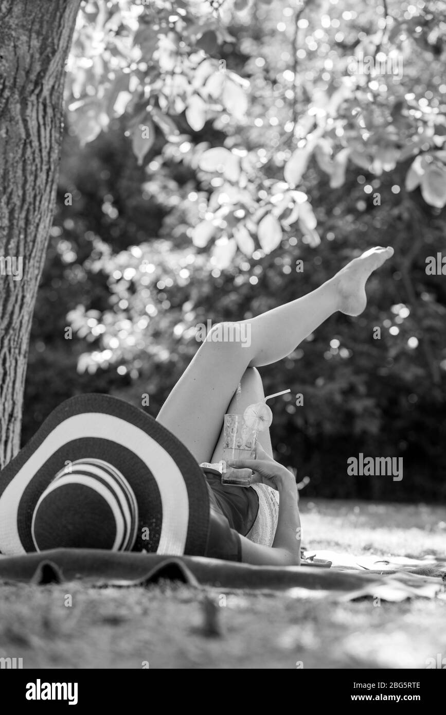 Monochrome image of a woman relaxing in the garden lying on a rug on the grass in her hat with a refreshing glass of water. Stock Photo