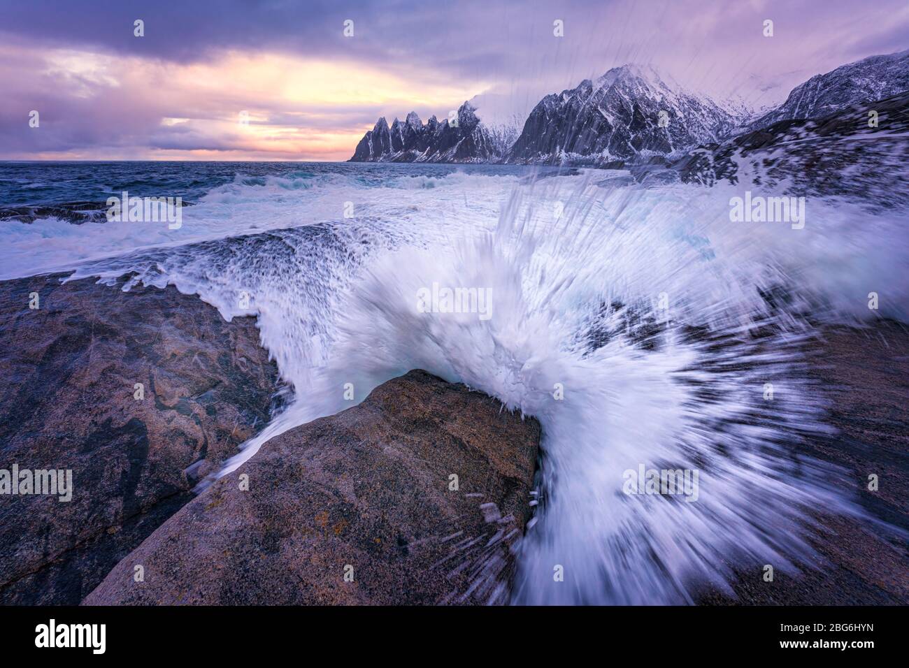 Huge waves crashing onto the coastal rocks at high tide in the morning, with colorful sky and jagged snow mountains on the horizon. Stock Photo