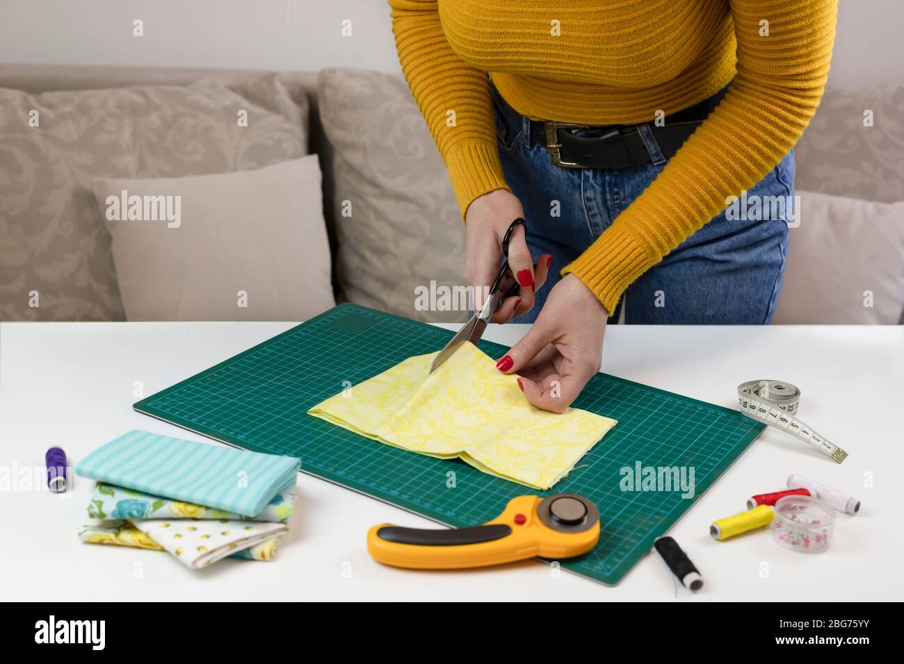 woman in a yellow cuts the fabric. there are quilting tools on the table. patchwork knife, scissors, lined cutting mat, self-locking, thread, centimet Stock Photo
