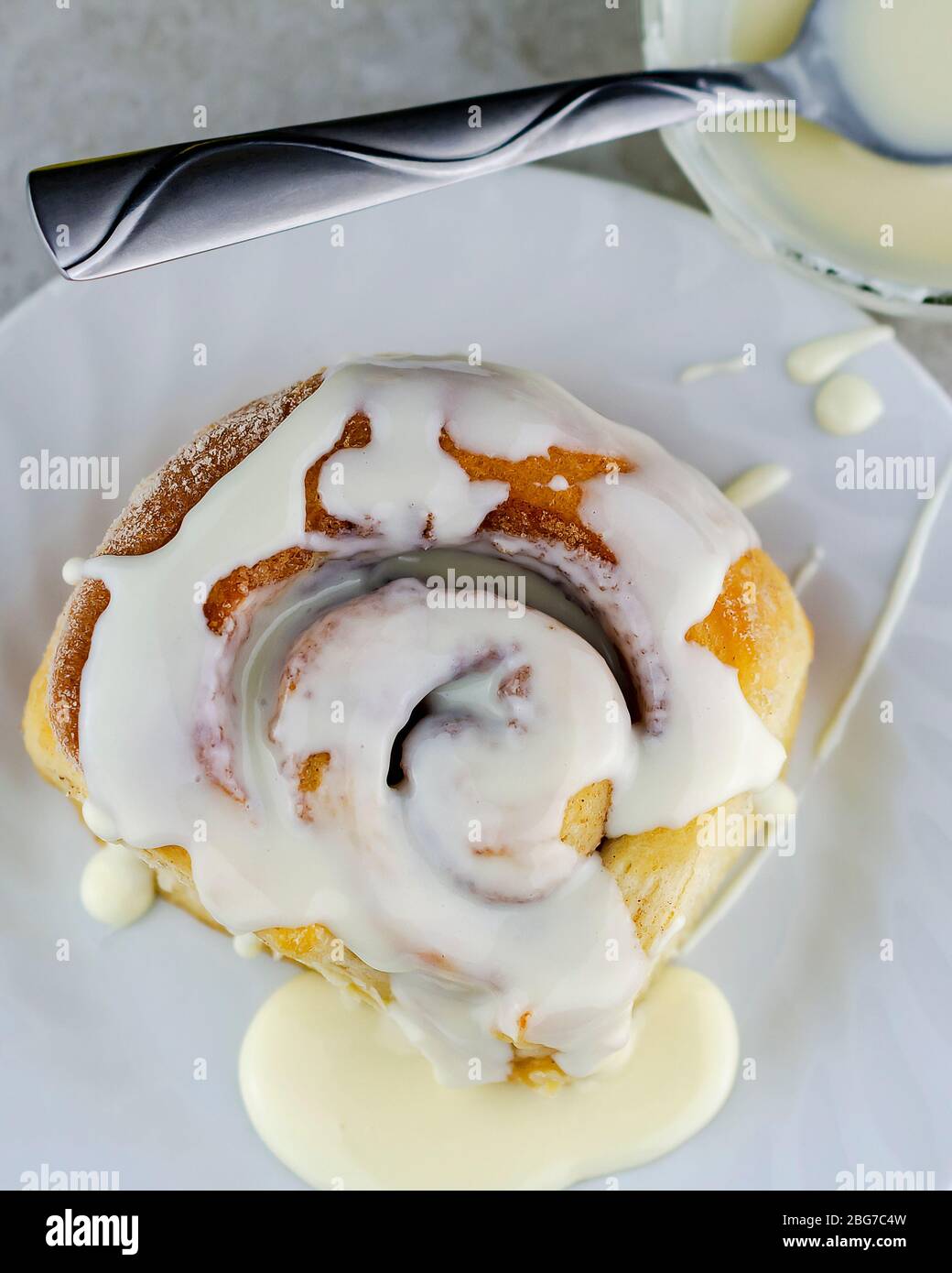 Overhead shot of a cinnamon roll on a white plate with a bowl of glaze. Stock Photo