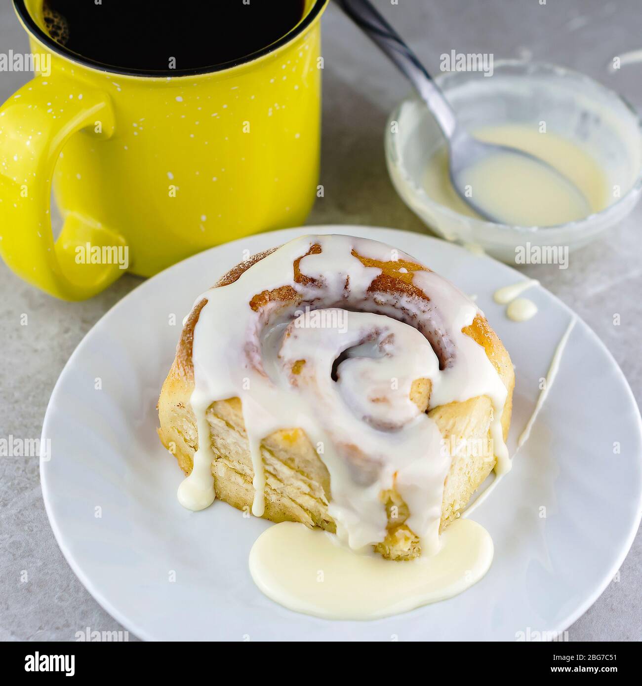 Cinnamon roll on a white plate with cup of coffee and bowl of glaze in the background. Stock Photo