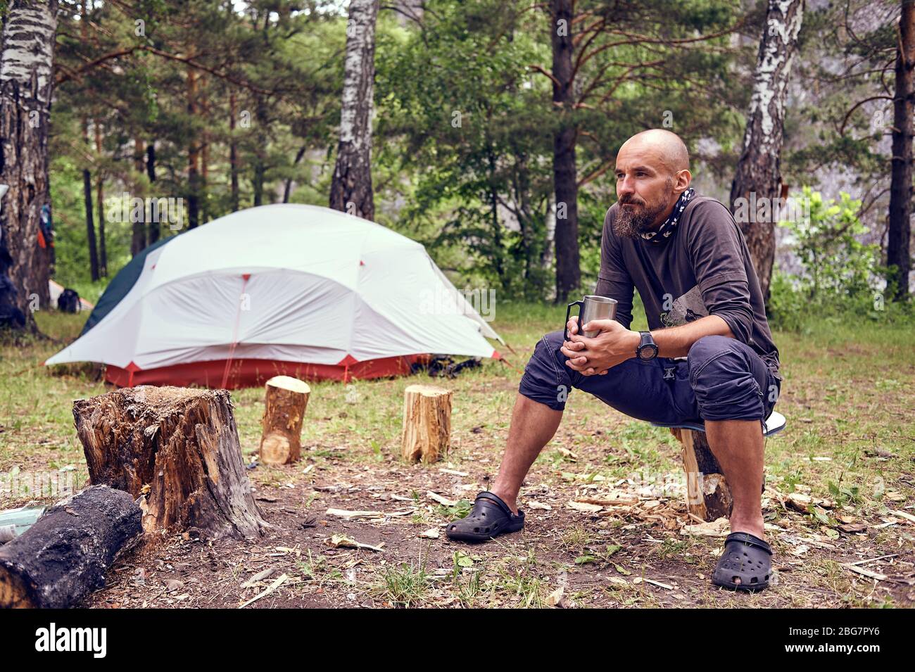 A young, bearded man drinks tea on a camping trip, in nature, against the backdrop of a tourist tent. Stock Photo
