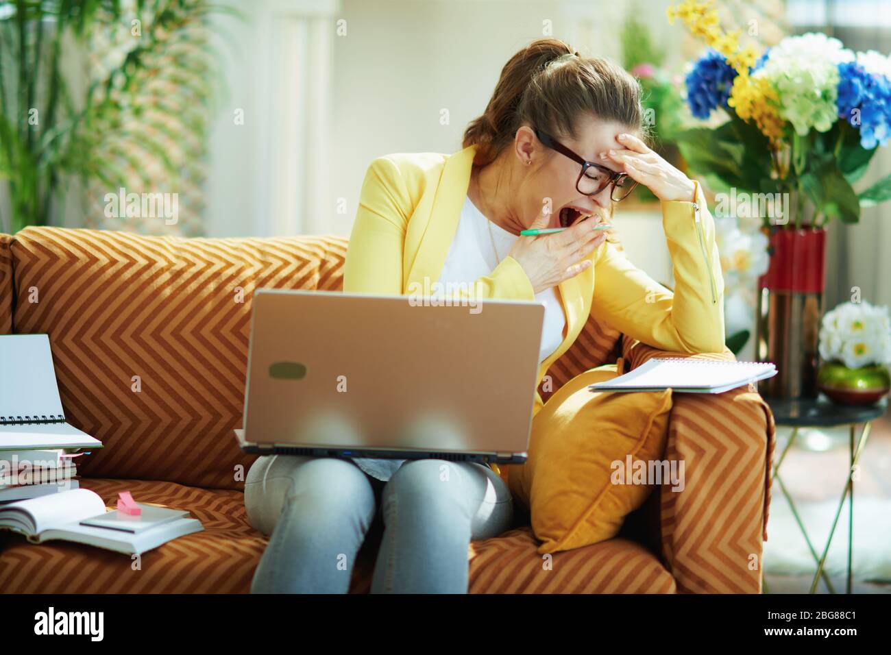 tired young housewife in jeans and yellow jacket in the modern living room in sunny day study online on a laptop. Stock Photo