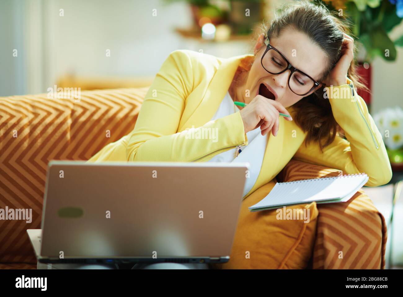 tired elegant student woman in jeans and yellow jacket in the modern house in sunny day study online on a laptop. Stock Photo