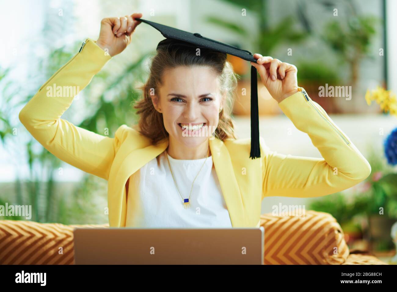Portrait of happy modern middle age housewife in jeans and yellow jacket with graduation cap and laptop in the modern house in sunny day. Stock Photo