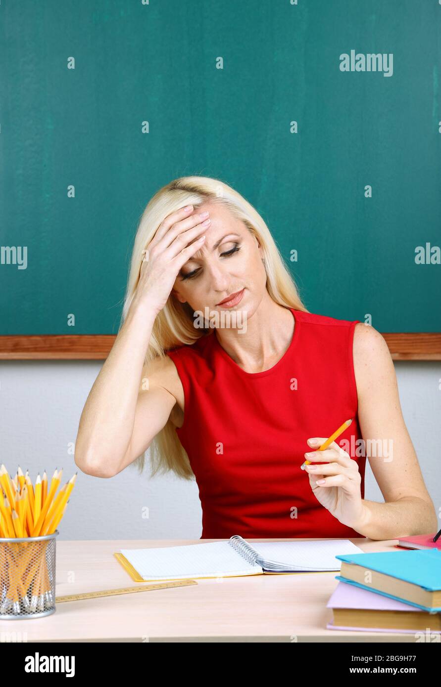 Tired teacher sitting at table on blackboard background Stock Photo