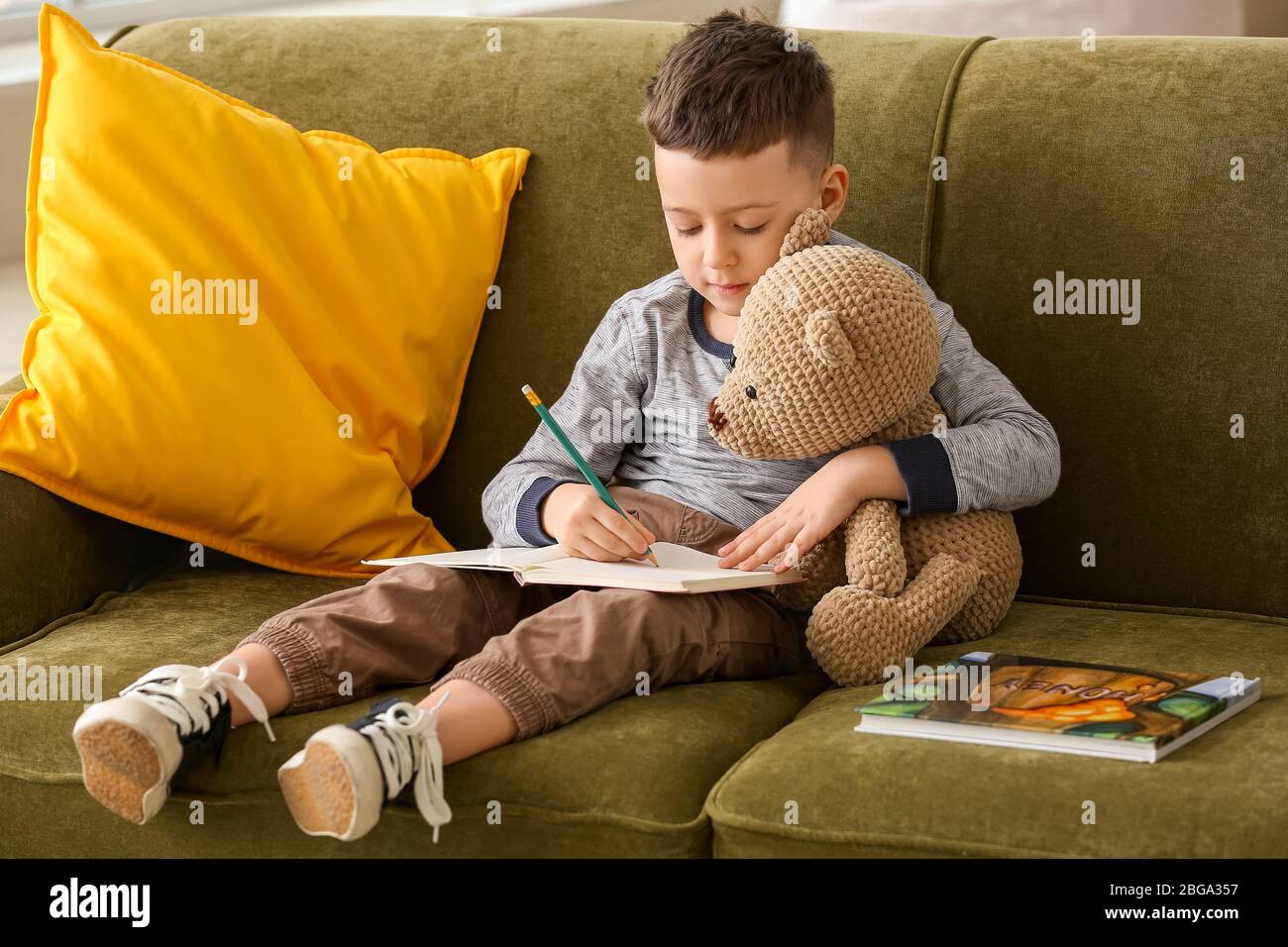 Cute little boy with teddy bear and notebook sitting on sofa at home ...