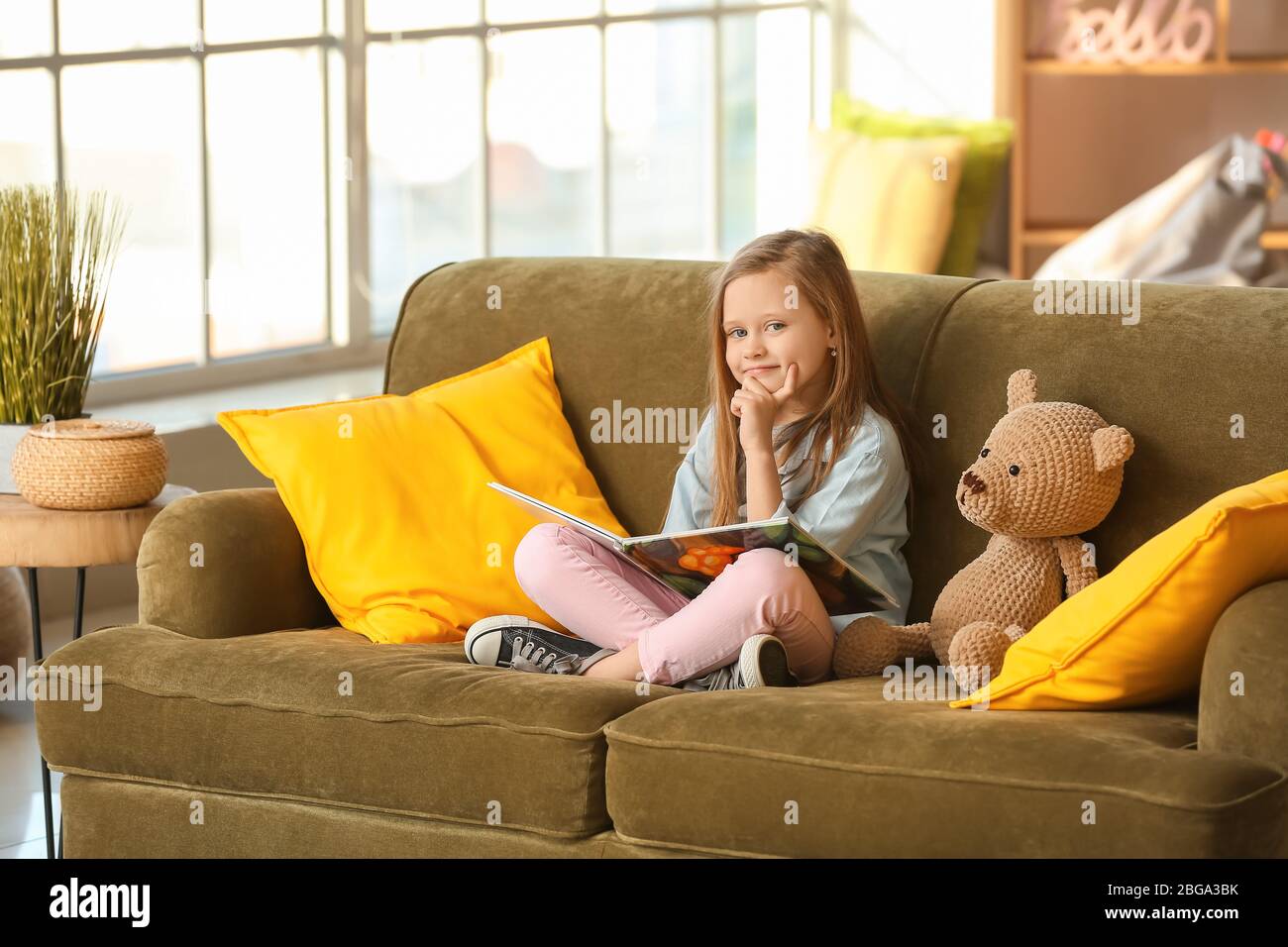 Cute little girl reading book on sofa at home Stock Photo - Alamy