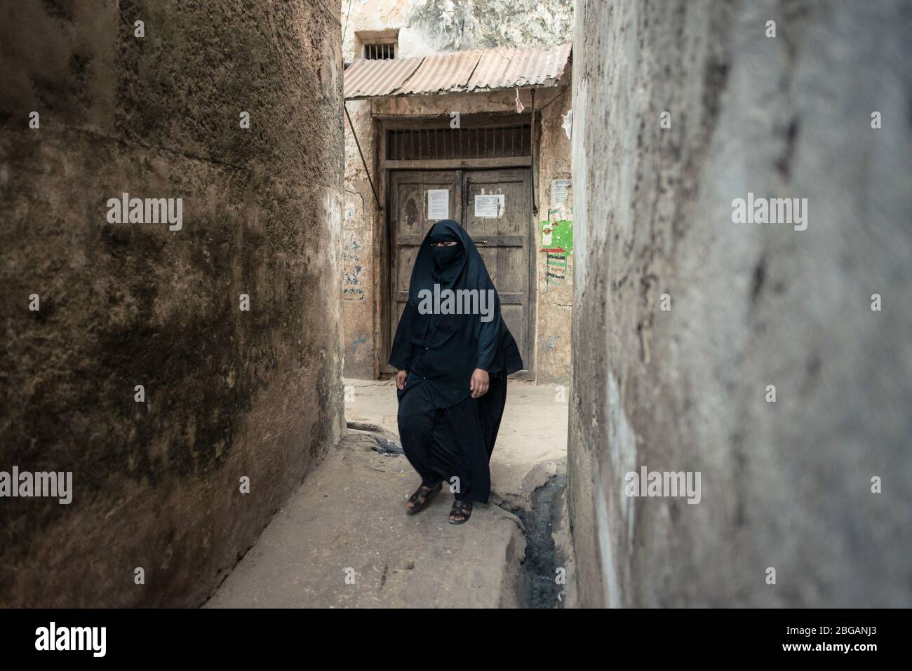 Adult Muslim woman wearing conservative niqab, only eyes visible, walking in narrow alley in Lamu island town, Kenya Stock Photo