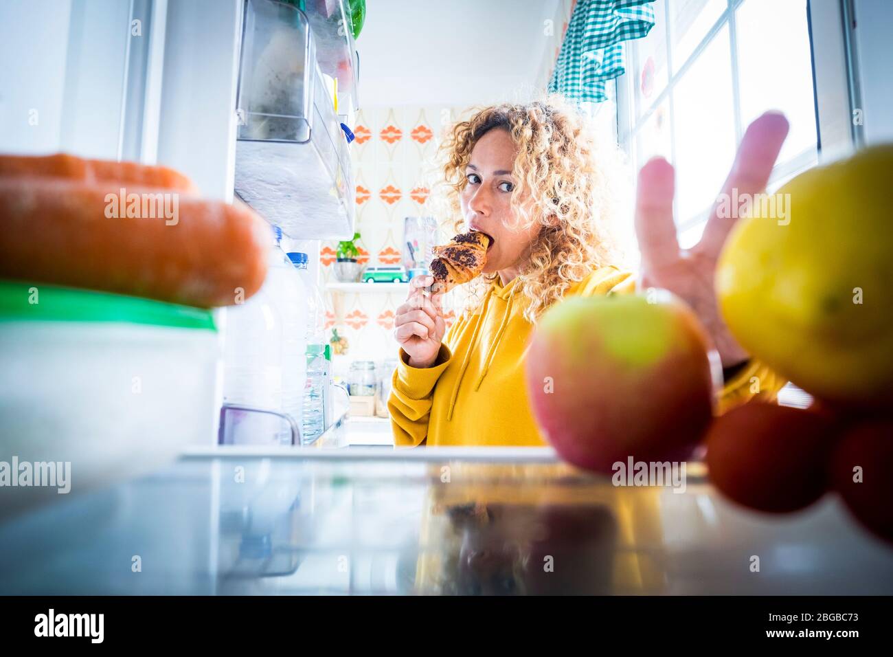 Alternaive inside view of Beautiful hungry adult woman eat tasty croissant with chocolate and open the fridge to take off some food -  concept of coro Stock Photo