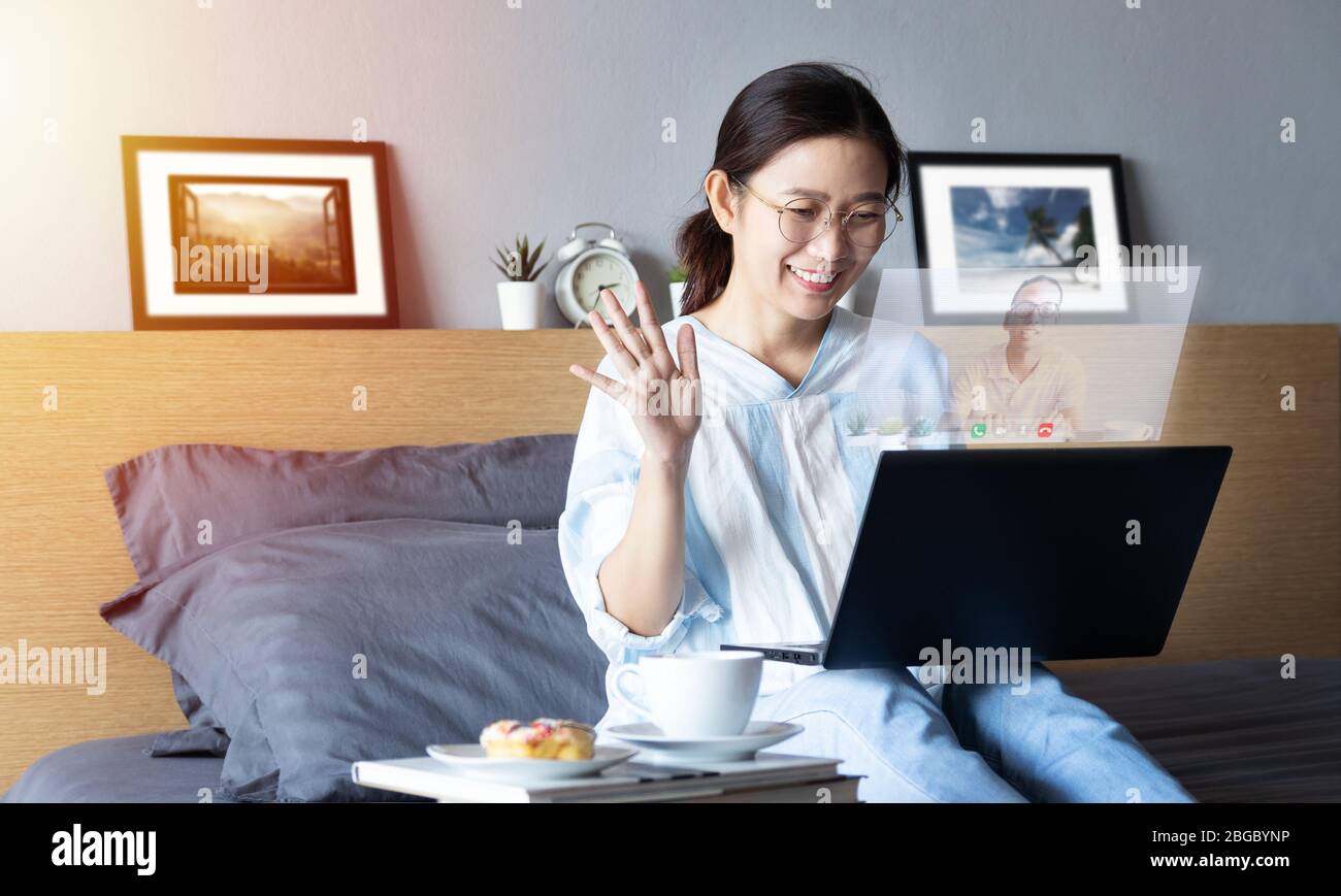 attractive happy woman using video call, make online conversation via laptop computer on bed from her room, having video chat, waving hand with visual Stock Photo