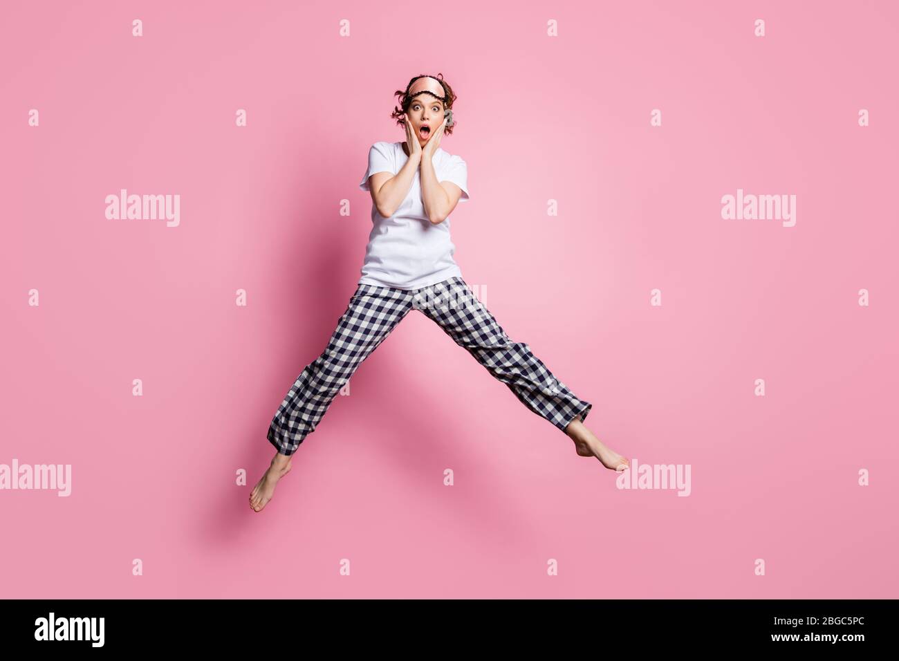 Full length photo of funny lady jump high up hands on cheeks open mouth wear sleep clothes mask white t-shirt plaid pajama pants barefoot isolated Stock Photo