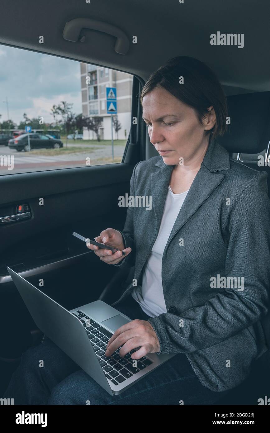 Businesswoman using mobile phone and laptop computer in car whole sitting at the back seat, business communication on the move Stock Photo