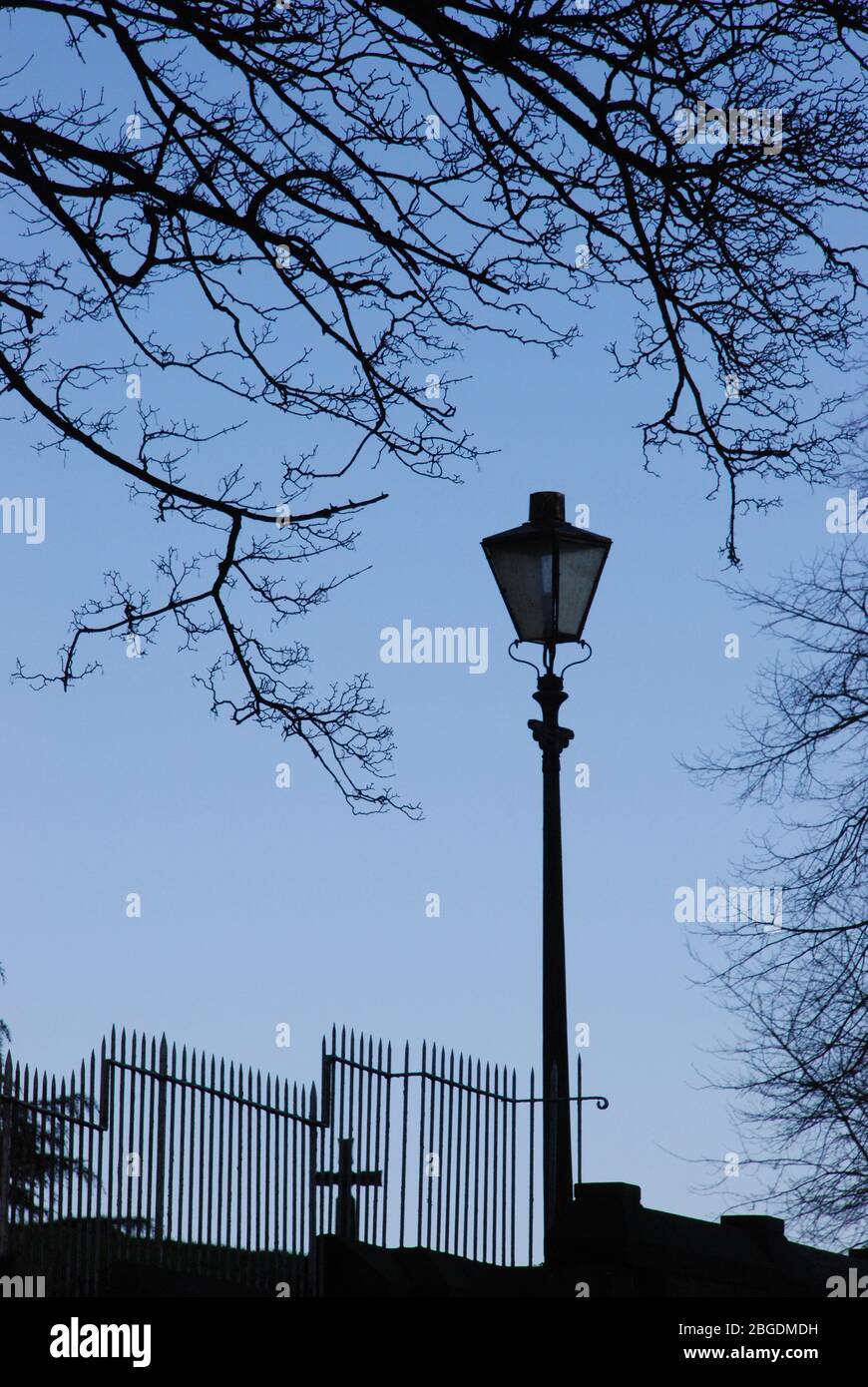 Single street light with wrought iron staggered fence and cross behind along with leafless tree curving around light all in silhouette Stock Photo