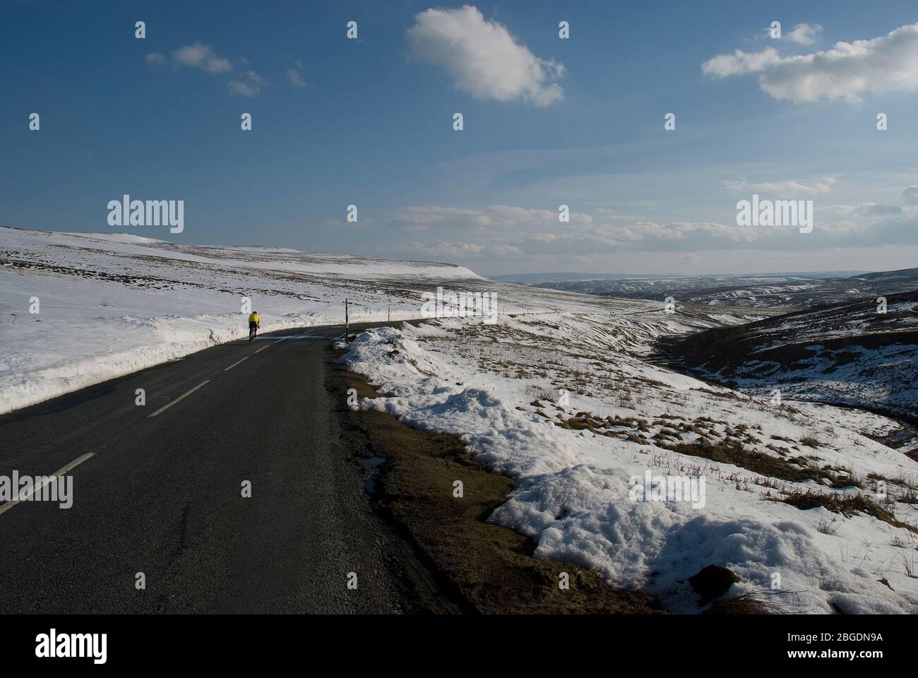 Lone cyclist wearing a yellow jersey cycling over a cleared moorland road in County Durham during winter with the landscape covered in snow Stock Photo