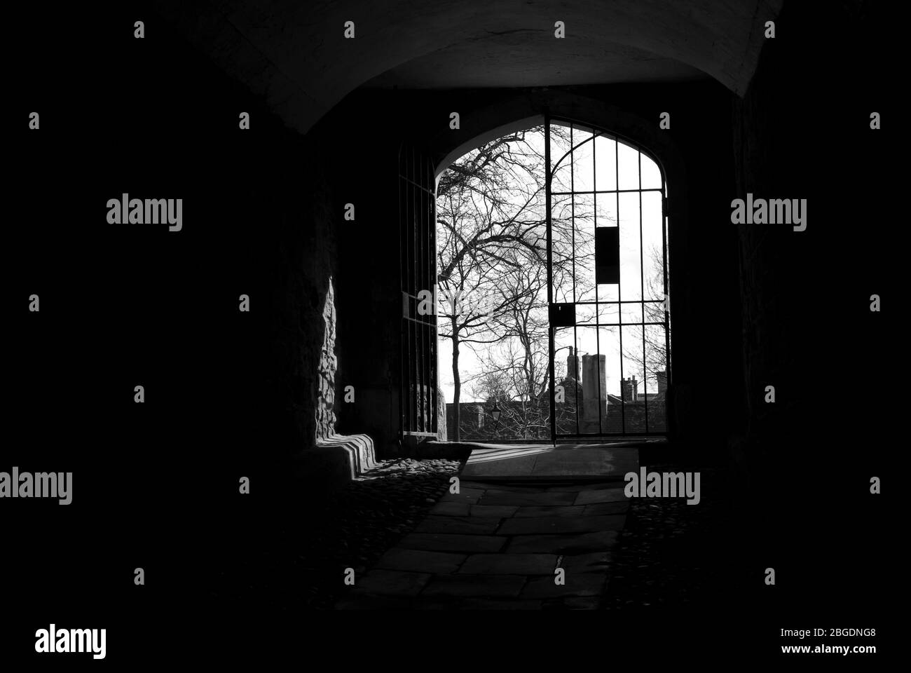 Monochrome image showing a dark passage way leading to a large metal gate set in the wall with one side open and sun coming in Stock Photo