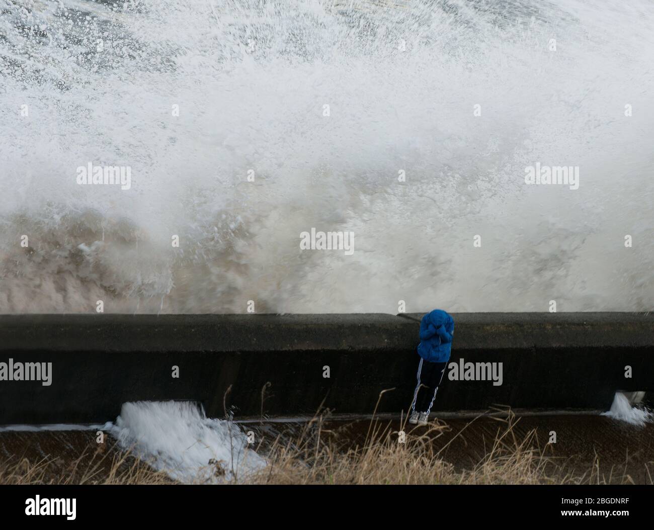 Figure of small individual covering face and head whilst standing behind promenade wall as stormy sea waves smash into and over the  wall Stock Photo