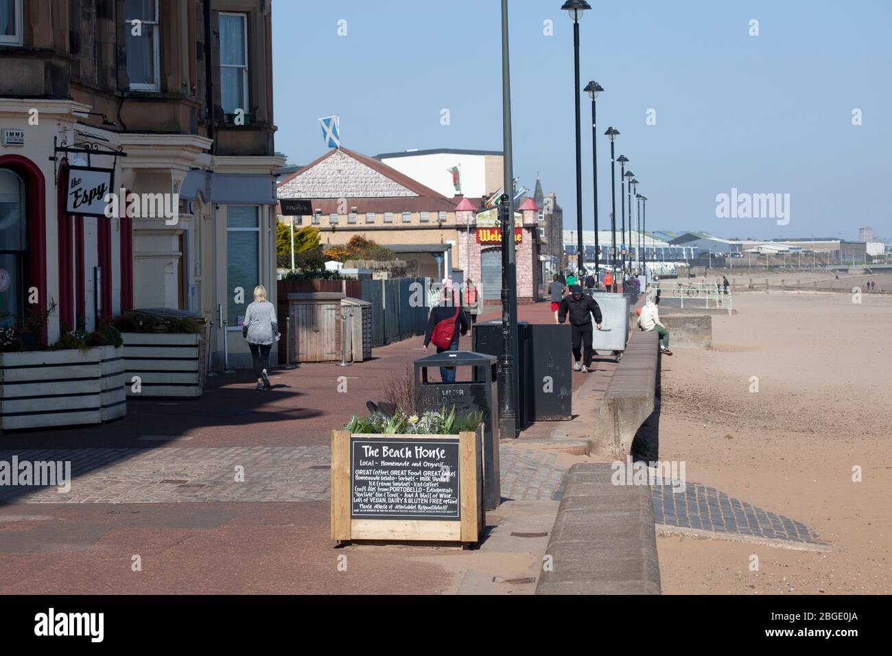 Portobello, Edinburgh, Scotland, UK. 21st April 2020. As Coronavirus Lockdown continues the public who are out before noon enjoy the sunshine at the seaside however, a cool ENE wind of 30km/h with potential gusts of 46 km/h does not encourage those that are out to sit for very long. Three places are now opening for take away food, Muro's Bistro, The  Beach House and the Espy Pub has removed the shutters from their windows. Credit: Arch White/ Alamy Live News. Stock Photo