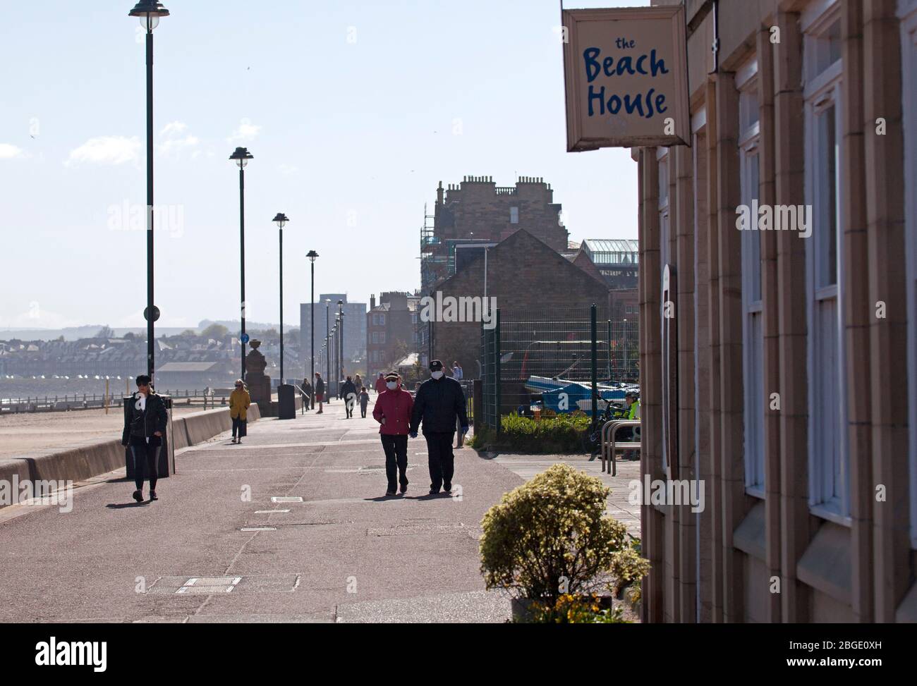 Portobello, Edinburgh, Scotland, UK. 21st April 2020. As Coronavirus Lockdown continues the public who are out before noon enjoy the sunshine at the seaside however, pictured one elderly couple wearing face masks for protection. A cool ENE wind of 30km/h with potential gusts of 46 km/h does not encourage those that are out to sit for very long. Three places are now opening for take away food, Muro's Bistro, The  Beach House and the Espy Pub has removed the shutters from their windows. Credit: Arch White/ Alamy Live News. Stock Photo