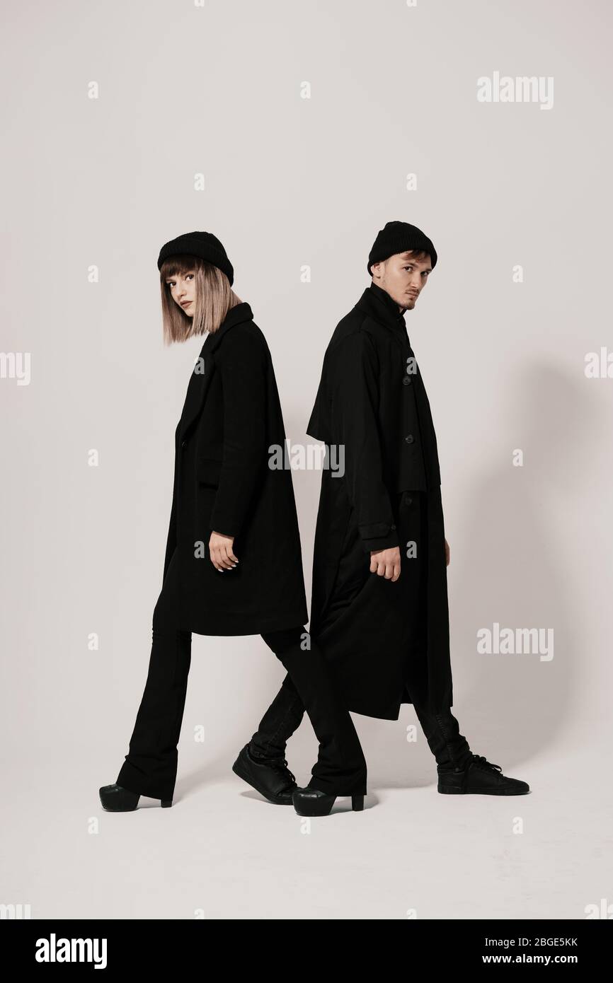 Full length side portrait of a Millenial young man and woman walking in different directions, wearing black clothes, hats on white background with cop Stock Photo