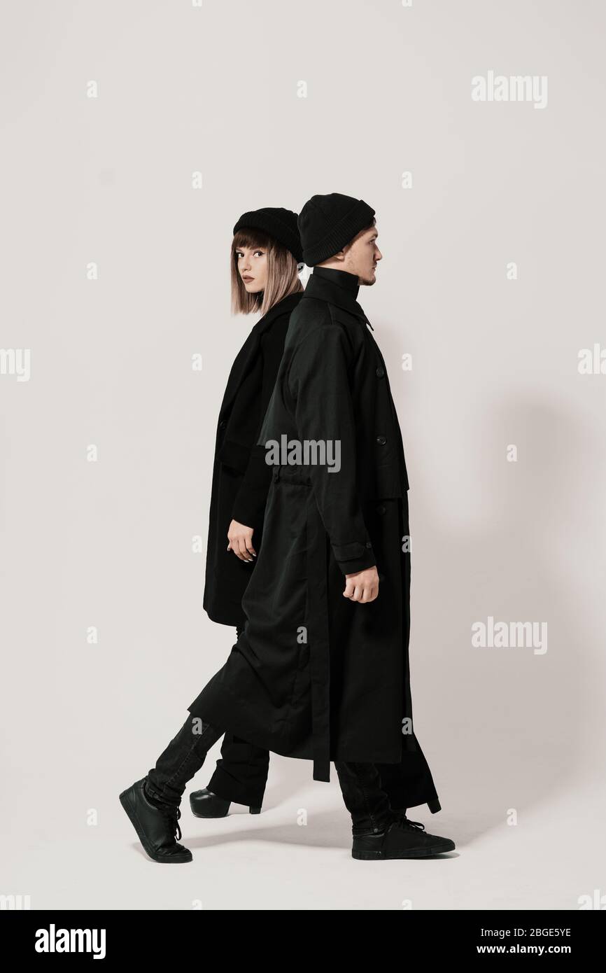 Full length side portrait of a Millenial young man and woman walking in different directions, wearing black clothes, hats on white background with cop Stock Photo